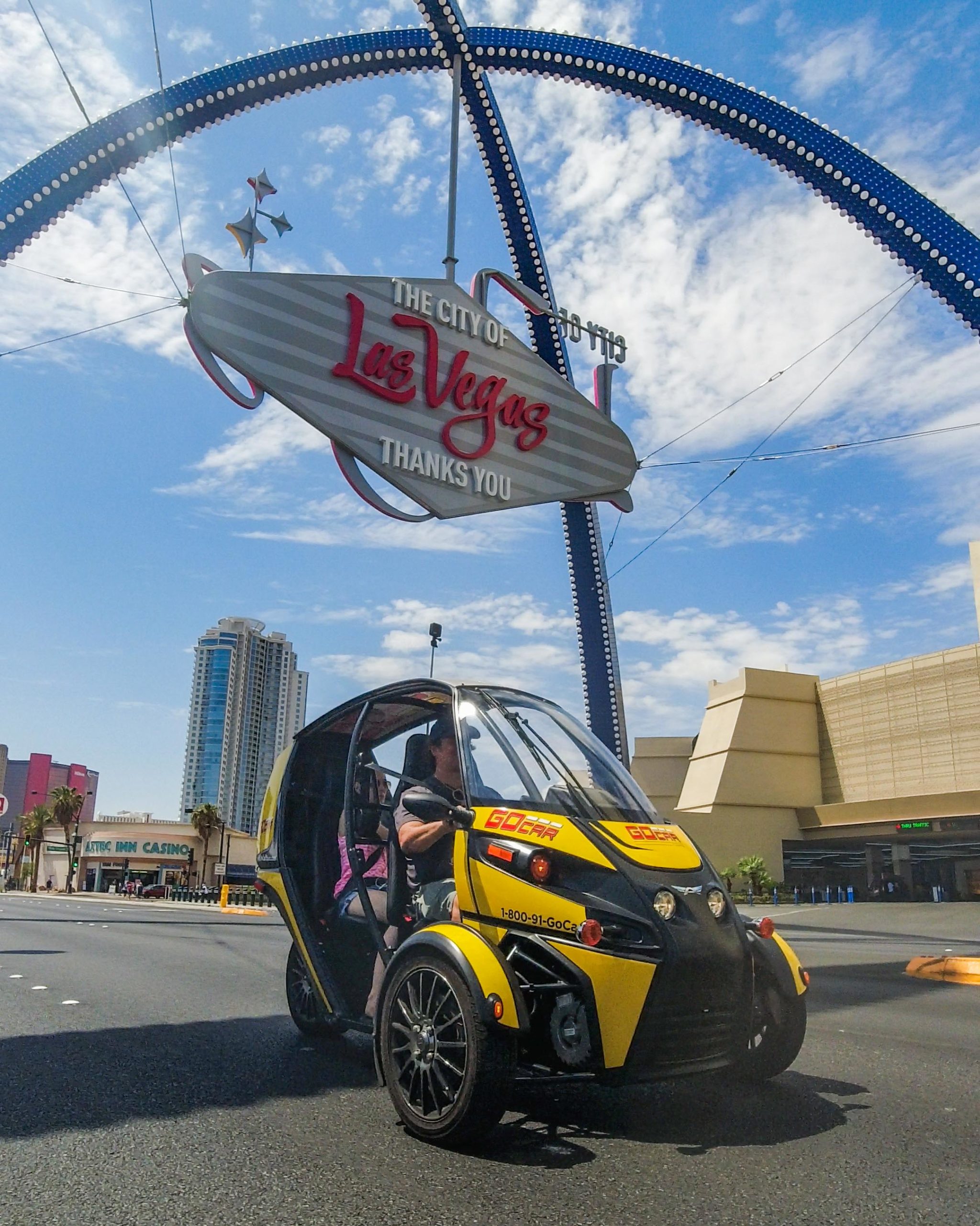 A yellow and black three-wheeled GoCar with a clear canopy roof, driving on Las Vegas Boulevard beneath the City of Las Vegas arch sign